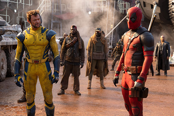 Wolverine in yellow and blue suit and Deadpool in red suit stand in a dusty outdoor setting.