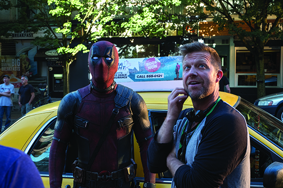 A person in a Deadpool costume stands next to a man in a black shirt by a yellow car.