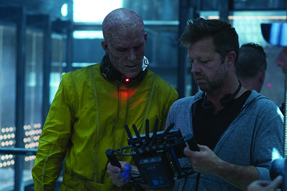 A man in bald prosthetic makeup and a yellow suit stands beside a crew member holding a monitor.