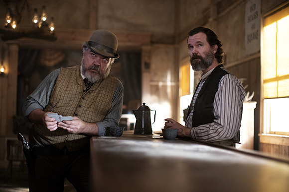 Two bearded men in 19th-century attire stand behind a wooden bar in a dimly lit room.