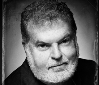 Black and white close-up portrait of a man with wavy hair and a full white beard looking forward.