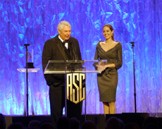 A man in a tuxedo and a woman in a grey dress stand behind an ASC podium on a blue lit stage.