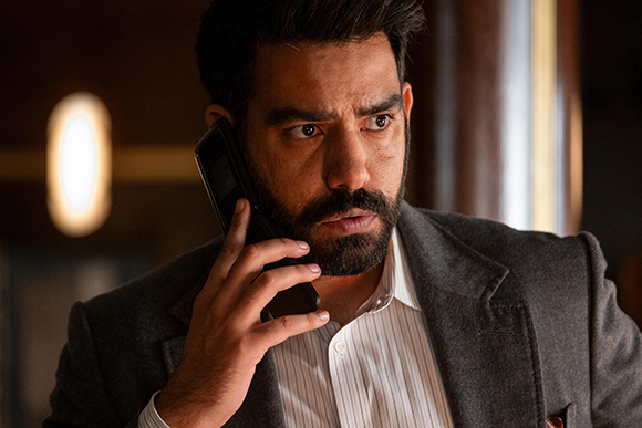 A man with dark hair and a beard talks on a black smartphone while wearing a grey blazer and striped shirt.
