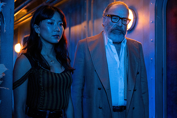 A young woman with dark hair and an older man with glasses and a beard stand in blue-toned lighting.