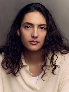 A woman with long, dark, wavy hair and a neutral expression looks directly at the camera.