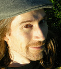 A man with long hair and a mustache wearing a grey cap, looking toward the camera.
