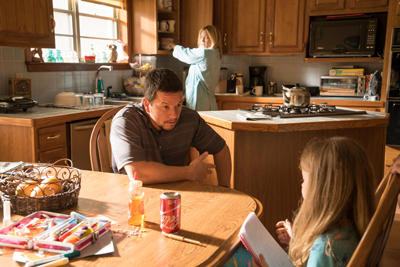 A man sits at a wooden table while a woman works in the background of a sunlit kitchen.