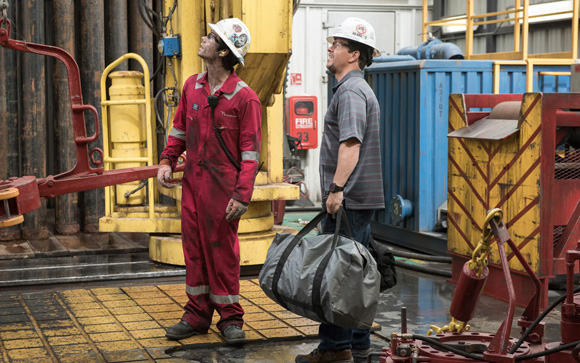 Two men in hard hats stand on a metal drilling rig floor amidst heavy machinery.