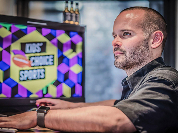A man with short hair and a beard sits at a desk looking toward a monitor displaying colorful graphics.