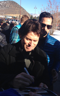 A man with brown hair and a black coat smiles while signing autographs for fans outdoors in the sun.