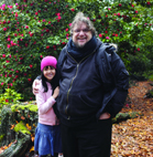 A man in a black jacket and a young girl in a pink hat smiling in front of red flowers.