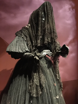 A mannequin wearing a black Victorian-era dress with a full skirt and a black lace veil covering its head and face. The backg