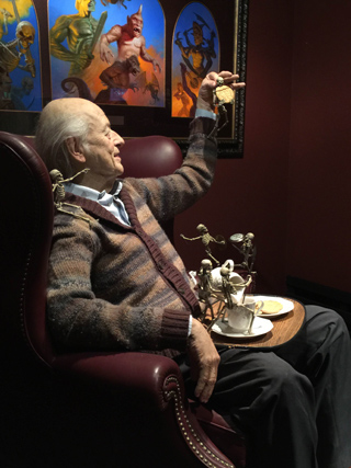 A wax figure of an elderly man in a cardigan sits in a leather chair, holding a medal, with small skeleton figures around him