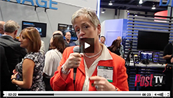 A woman in a red blazer holds a microphone while speaking at a busy indoor convention.