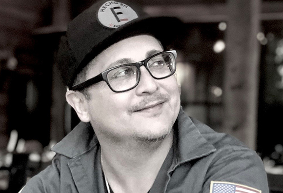 Black and white portrait of a man wearing a baseball cap and glasses, looking upward and smiling slightly.