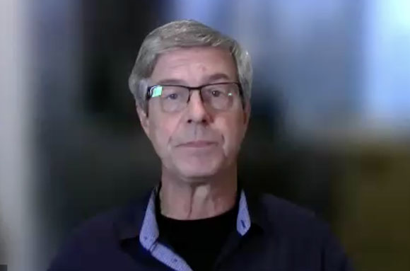 A man with gray hair and glasses wearing a dark blue button-down shirt over a black shirt.