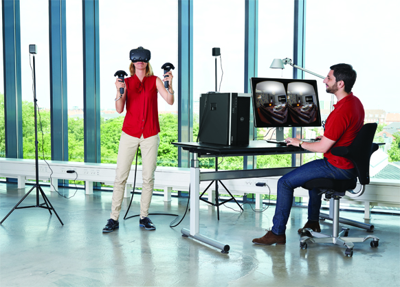 A woman in a red shirt uses a VR headset and controllers while a man sits at a desk monitoring a computer screen.