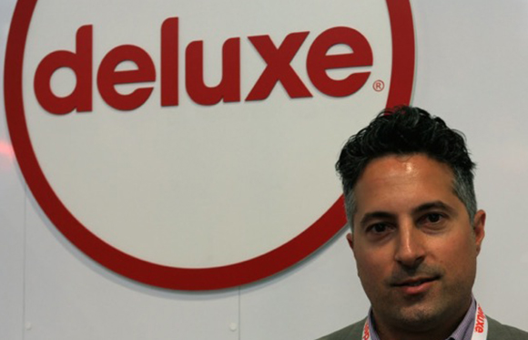 A man with dark hair looking toward the camera in front of a large red and white Deluxe logo.