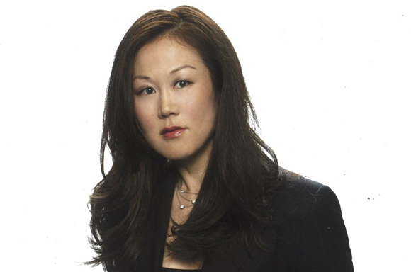 A woman with long dark hair and a black blazer looks toward the camera against a white background.