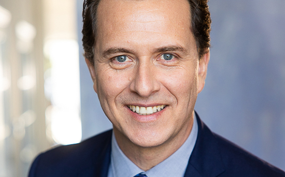 A smiling man with brown hair wearing a blue suit jacket and light blue shirt.