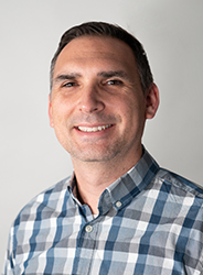 A man with short dark hair smiling while wearing a blue and white plaid button-down shirt.