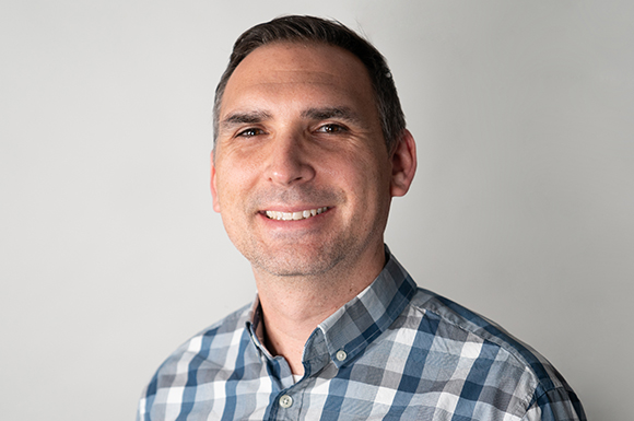 A man with short dark hair and light stubble wearing a blue and white plaid button-down shirt.