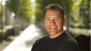 A smiling man with short brown hair wearing a black shirt, positioned against a blurred outdoor background.