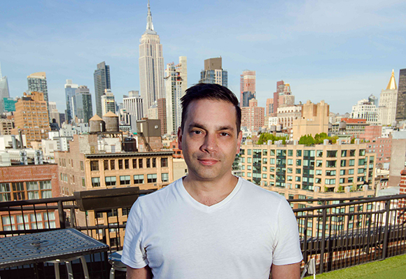 A man in a white t-shirt stands on a rooftop with the Empire State Building and NYC skyline behind him.