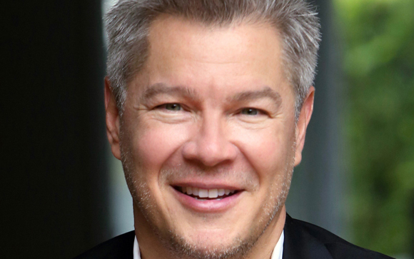 A close-up portrait of a smiling man with short gray hair and light stubble.