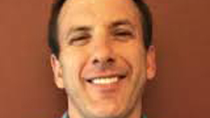 A man with short dark hair smiling broadly against a solid reddish-brown background.