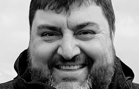 Black and white close-up of a smiling man with dark hair and a full beard.