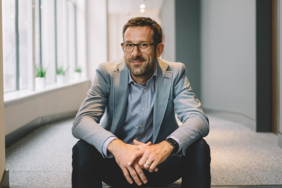 A man with glasses and a beard wearing a grey blazer sits on a carpeted floor in a hallway.