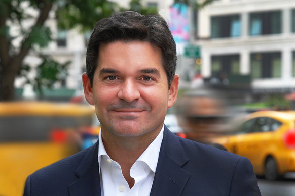 A man with dark hair and a slight smile wearing a navy blazer and white collared shirt.