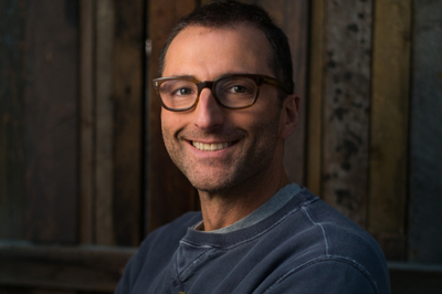 A smiling man with short dark hair and brown-rimmed glasses wearing a dark blue sweatshirt.