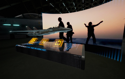 A digital control panel with playback icons in front of a large curved screen showing silhouettes at sunset.
