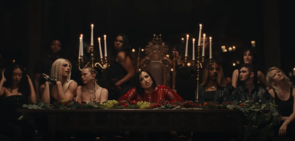 A group of people in dark clothing sit around a long table decorated with fruit and greenery under candlelight.