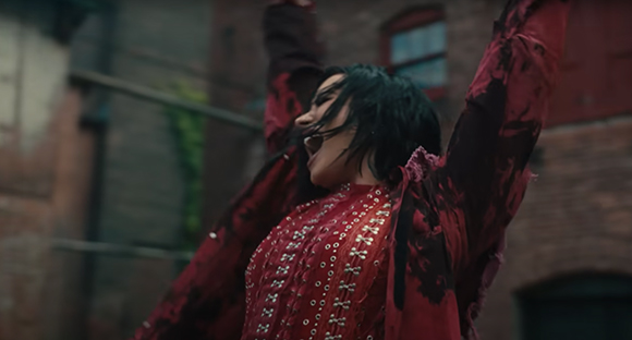A person with dark hair wearing a red patterned top and jacket, arms raised against a brick building.
