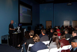 A presenter at a screen addresses an audience seated in a dark room.