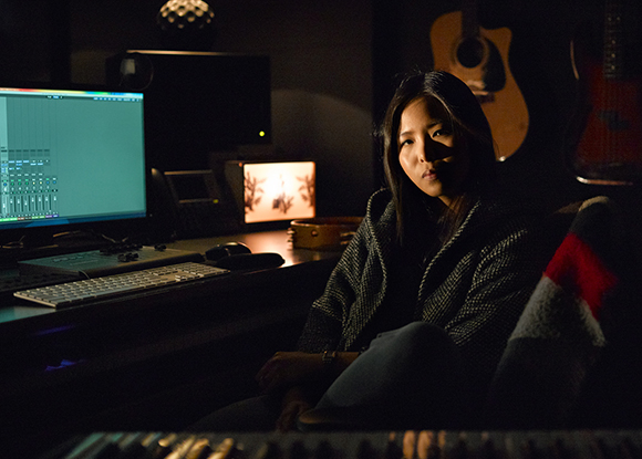 A woman sits in a dark music studio next to a computer monitor, keyboard, and guitars.