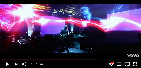 A person plays guitar on a dark stage amidst flowing red and white light streaks and large video projections.