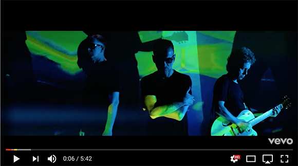 Three men perform on a stage illuminated by green and blue light projections.