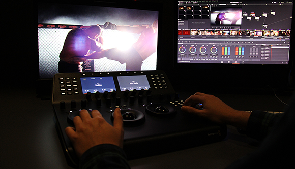 Hands operating a color grading console in front of dual monitors displaying video footage and editing software.