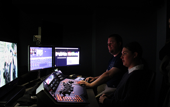 Two people sit in a dark room operating a video editing console with several glowing computer monitors.