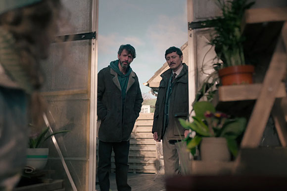 Two men stand in an open doorway, looking toward the viewer amidst potted plants and wooden structures.