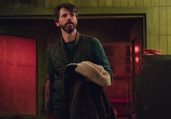 A bearded man in a green sweater and quilted vest looks up with a surprised expression while holding a coat.