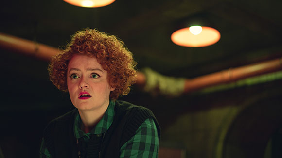 A woman with curly red hair and a surprised expression looks upward in a dimly lit setting.