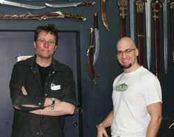 Two men pose in front of a dark wall decorated with various swords and daggers.