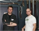 Two men pose in front of a wall display of various swords and blades.