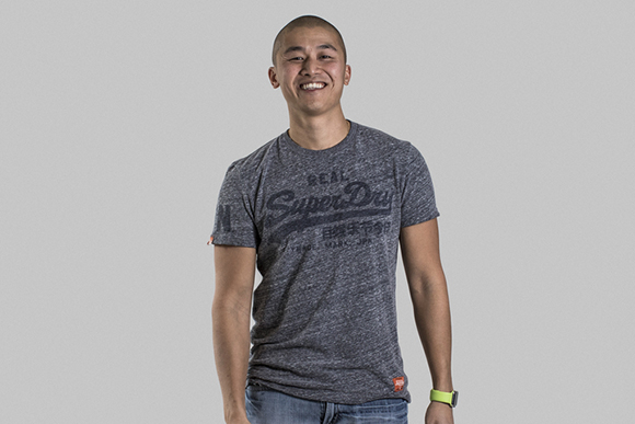 A smiling man wearing a grey graphic t-shirt and blue jeans against a grey background.