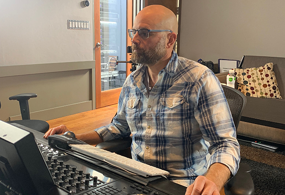 A bald man with a beard and glasses wearing a plaid shirt sits at a professional audio or video mixing console.
